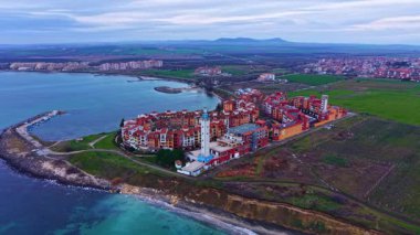 Deniz kıyısında birden fazla binası olan bir kasaba. Şehrin arkasında bulutlu bir gökyüzünün altında yeşil alanlar uzanıyor. Kıyı şeridi boyunca kıvrımlar var..