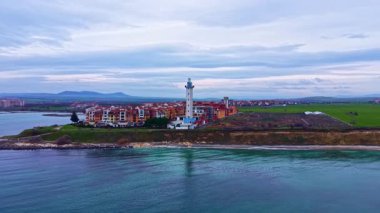 Bir deniz manzarası kıyı boyunca bir grup evin yanında duran bir deniz fenerini gösteriyor. Su sakin ve yeşil alanlar bulutlu bir gökyüzünün altında binaların arkasında uzanıyor..