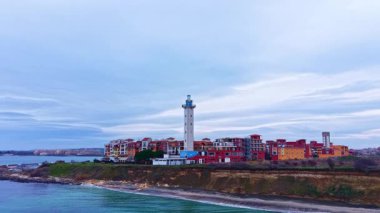 Renkli binaların yanında uzun bir deniz feneri duruyor. Kıyı şeridi akşam erken saatlerde bulutlu bir gökyüzü altında sakin dalgalarla karşılaşıyor..