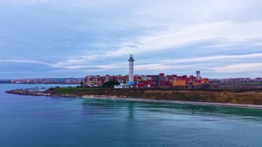 Bir deniz feneri kıyı boyunca bir grup binanın yanında duruyor. Su durgun, gökyüzünü yansıtıyor. Gün boyunca bulutlar havada sürüklenirken..