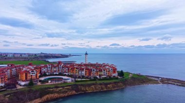 Arka planda deniz feneri olan deniz kıyısında bir tatil köyü var. Sahne geniş bir gökyüzü yakalıyor ve gri bulutların altında sakin bir deniz manzarası yaratıyor..