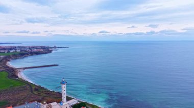 Okyanus manzaralı ve uzakta küçük bir kasaba olan bir deniz feneri kıyıya yakın durur. Bulutlar sakin suyun üzerindeki gökyüzünü kaplıyor..