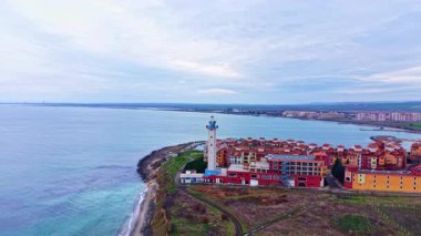 Yakınlarda binaları olan deniz feneri kıyıda duruyor. Kıyıya dalgalar vururken deniz kenarı kasabasında bulutlu bir gökyüzü beliriyor. Kıyı şeridi ufuk boyunca uzanıyor..