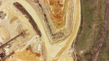 İnşaat makineleri bir kazı alanında çalışır, toprağı hareket ettirir ve araziyi şekillendirir. Zemin kum ve kil çizgileriyle işaretlenmiştir. Bölgenin kenarında ağaçlar görülebilir..