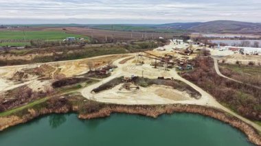 Makineler, kırsal bir alanda su birikintisine yakın bir inşaat alanında çalışıyor. Toprak temizleniyor ve gökyüzü bulutlu. Arka planda tarlalar ve tepeler görünür..