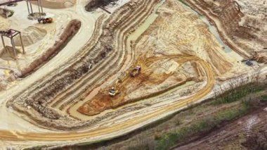 Ağır makineler inşaat alanında çalışıyor, toprağı hareket ettiriyor ve bir çakıl ocağında toprağı şekillendiriyor. Bölge toprak ve suyla dolu. Toprakta birden fazla iğne izi var..