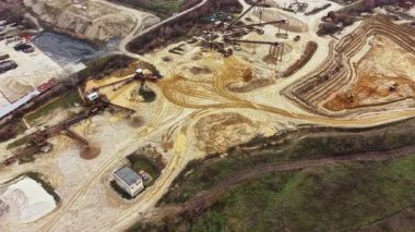 Makineler ve kamyonlar toprak ve çakıllarla dolu bir maden sahasında çalışıyor. Zemin, ekipman ve yapılar arasında toprak yığınları ve izler gösteriyor. İş gün boyunca devam eder..