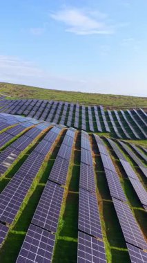 Güneş panelleri bir tepenin yamacına yeşil çimlerle çevrili. Güneş parlak mavi bir gökyüzünde parlıyor. Alan yenilenebilir enerjiye odaklandığını gösteriyor..