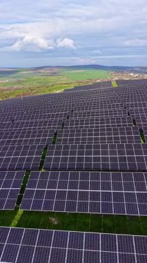 Güneş panelleri, arka planda yeşil çimenler ve tepeler olan bir alan boyunca uzanıyor. Gökyüzü birkaç bulutla mavidir. Güneş ışığı gündüz manzarayı aydınlatır..
