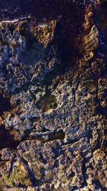 Kaya oluşumları kıyı boyunca görülebiliyor, gelgit çekildikçe taşların ayrıntıları ve dokuları ortaya çıkıyor. Güneş arka planda batarken su parıldıyor..