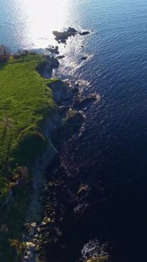 Sahil şeridi yeşil çimenler ve kayaların güneş ışığı altında berrak mavi suya değdiğini gösteriyor. Sahne, öğleden sonra kıyı boyunca karayı ve denizi aydınlatarak doğayı yakalar..