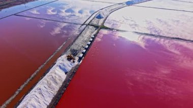Çarpıcı bir hava manzarası kırmızı ve pembe tonlarda canlı tuz düzlükleri yakalar, işçiler tuz toplarken bulutlar su yüzeyine yansıyarak nefes kesici bir manzara yaratır..