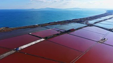 Sersemletici bir hava manzarası, berrak bir gökyüzü altında sakin sular ve uzak dağlarla güzel tezat oluşturan, kırmızı ve mavi tonlarda geniş tuz düzlükleri sergiliyor..