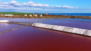 Göz kamaştırıcı bir tuz tavası görüntüsü gökyüzünü yansıtan canlı kırmızı renkler ile gözü yakalar. İşçiler özenle tuz toplarken manzaralı evler yeşil araziyi dolduruyor..