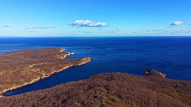 Kıyı şeridi kayalık alanlar ve temiz su karışımını gösteriyor. Toprak çoğunlukla ağaçlar ve bitkiler olmadan çıplak ve bulutlar mavi gökyüzüne dağılmış durumda..