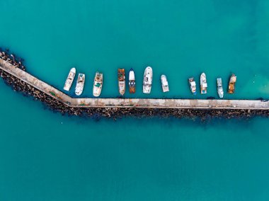 Tekneler taş bir iskele boyunca bir sıra halinde rıhtımda görülürler. Su berrak ve mavidir, gün ışığında çevreyi gösterir. Sahne huzurlu kıyı manzarasını yakalıyor, hiç insan yok..