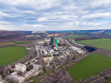 Bir çimento fabrikası yeşil tarlalar ve tepelerle çevrili işçiler ve makinelerle meşgul. Gökyüzü bulutlu ve manzarada yakınlarda küçük bir kasaba var..
