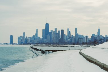 Arka planda Chicago silueti olan karlı bir yürüyüş yolu rıhtım boyunca uzanıyor. Binalar, tipik kış havası gibi gri gökyüzüne yükselir..