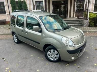 Küçük bir minibüs bir binanın önündeki taş bir yolda park edilmiş. Çevrede ağaçlar var, ve bazı yapraklar yerde, sonbaharın başlarını gösteriyor..