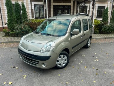 Modern bir binanın önüne düzgün park edilmiş gümüş bir minibüs. Bölgede ağaçlar ve çiçekler var ve hava açık. Minibüste hasar yok..