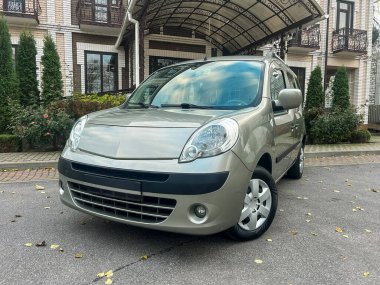 Tuğla görünümlü büyük bir binanın önünde küçük bir araba park edilir. Bölge ağaçlarla çevrili ve bakımlı görünüyor. Gökyüzü açık..