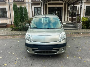 Büyük bir binanın önünde gümüş renkli bir araba park edilmiş. Her iki tarafta da yeşil bitkiler ve girişe giden merdivenler var. Gündüz oldu ve gökyüzü açık..