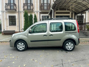 Açık renkli bir binanın yanında gümüş renkli bir araç park edilmiş. Arka planda ağaçlar ve tepe örtüsü var. Görüntü, etrafta kimsenin olmadığı açık bir günü gösteriyor..