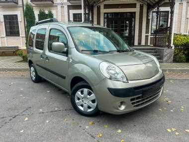 Çevresinde taştan bir veranda ve yeşil bitkiler olan bir binanın önündeki asfalt bir alana park edilmiş gümüş bir minibüs var. Hava açık ve açık..