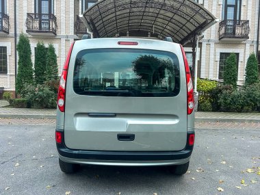 Klasik tasarımı olan bir binanın önüne gümüş bir minibüs park edilmiş. Minibüsün görünürde kimse olmadan basit bir dikiz açısı var. Yakınlarda yeşil ağaçlar görülüyor..