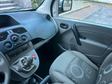 Küçük bir aracın ön tarafında gösterge paneli, vites kolu ve koltuklar var. Manzara ön cama ve yan aynaya bakıyor. Dışarıda bir sokak ve ağaçlar görünüyor..