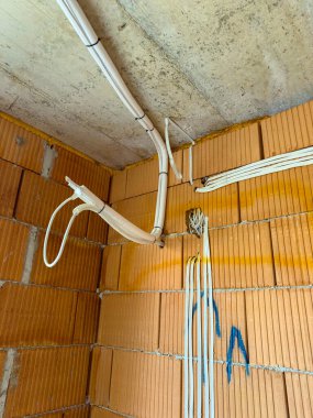 İşçiler inşaat halindeki bir odanın köşesine elektrik kabloları ve borular kuruyorlar. Duvarlar turuncu tuğlalardan yapılmış ve tavan tamamlanmamış..