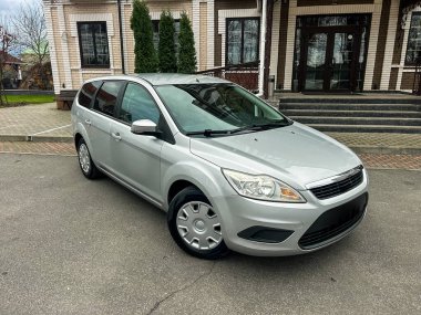 Şehir merkezindeki bir binanın önünde gümüş renkli bir araba park halinde. Araç pürüzsüz bir yüzeyde tek başına duruyor ve hiç insan yok. Arka planda yeşil bitkiler var.