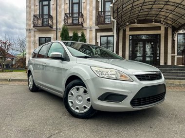 Büyük bir binanın önünde gümüş renkli bir araba park edilmiş. Hava bulutlu ve ortam kentsel. Araba kameraya bakıyor ve net detaylar gösteriyor..