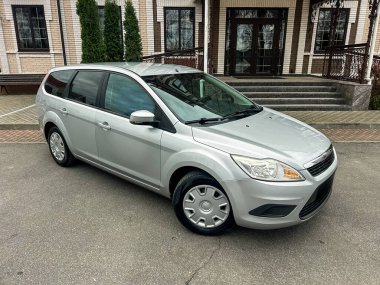 Gümüş renkli bir araba bir binanın önündeki kaldırımda park halinde. Binanın büyük pencereleri ve girişe çıkan merdivenleri var. Yakınlarda yeşil ağaçlar var..