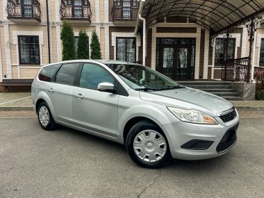 Gümüş renkli bir araba tuğla duvarları ve birçok penceresi olan bir binanın yanında park edilmiştir. Hava bulutlu. Ayarlar yerleşim alanı gibi görünüyor..