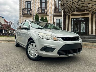 Büyük bir binanın önündeki bir yolda gümüş bir araba park edilmiştir. Binanın birden fazla penceresi ve merdiveni var. Gökyüzü bulutlu, bu da yağmur yağabileceğini gösteriyor..