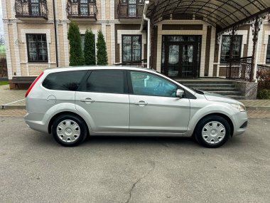 Büyük bir binanın önünde yolun kenarında gümüş renkli bir minivan park edilmiş. Araç, zemininde yeşil ağaçlar olan asfalt bir yüzeye yerleştirilmiş..