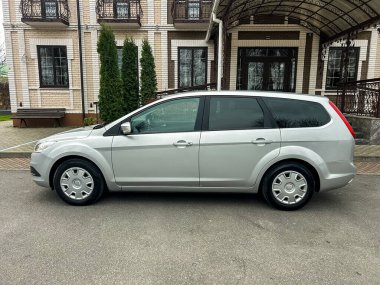 Büyük bir konut binasının önündeki asfaltta gümüş bir araba park halinde. Binanın birkaç katı var ve çevresi yeşil bitkilerle çevrili. Gündüz oldu..