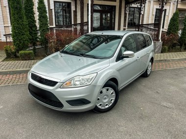 Gümüş renkli bir araba yolun kenarında, bir binanın yanında park halinde. Ortam ağaçlar ve temiz yollar içeriyor. Gün ışığı arabanın tasarımının detaylarını gösteriyor.