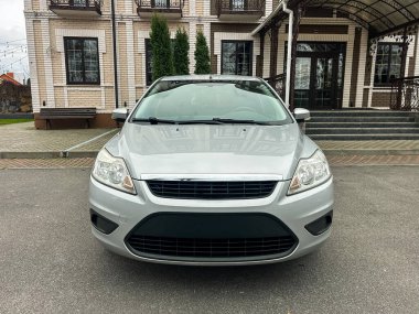 Büyük bir binanın önünde gümüş bir araba park edilmiş. Araba izleyiciye bakıyor. Patikada taş fayanslar var. Gökyüzü gri ve bulutlu görünüyor. Ağaçlar yakınlarda görünüyor..