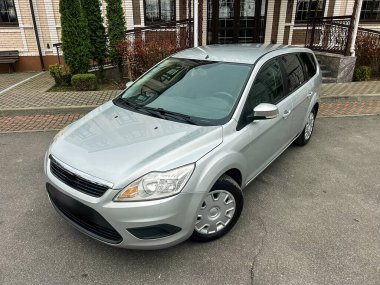 Gümüş renkli bir araç bir binanın yakınındaki asfalt bir yüzeye park edilmiş. Arka planda yeşil ağaçlar ve açık bir gökyüzü var. Sahne dışarıdaki tipik bir günü gösteriyor..