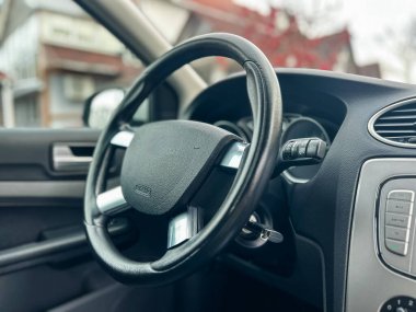 Direksiyon ve gösterge paneli park halindeki bir arabanın içinde görünür. Kontrol düğmeleri direksiyonun yanında. Parlak güneş ışığı pencerelerden görünür..