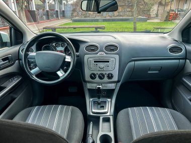 Arabanın içi gösterge paneli, direksiyon simidi ve koltukları gösteriyor. Dışarıda yeşil alanlar ve gün boyunca sakin bir yerde bir taş bina var..