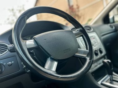 Arabanın içi direksiyon, torpido gözü ve vites kolunu gösteriyor. Araç bir binanın yanında park halinde. Ayar gündüz vakti, arabanın içinde kimse yok..