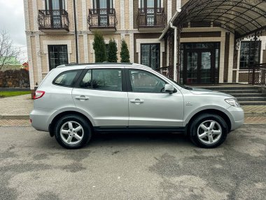 Gümüş renkli bir SUV tuğla duvarlı ve birden fazla balkonlu büyük bir binanın yanına park edilmiş. Zemin asfalt kaplı. Gökyüzü bulutlu, bu da yağmur yağabileceğini gösteriyor..