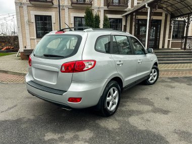 Gümüş renkli bir SUV büyük bir tuğla binanın yanında park edilir. Çevrede açık bir garaj yolu ve yeşil ağaçlar var. Sahne bulutlu bir günde, tipik bir banliyö bölgesinde geçiyor..