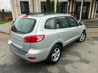 Gümüş bir araba bir binanın yakınındaki düz bir yüzeye park edilmiştir. Arabanın çatı rafı var ve durumu iyi. Arabanın çevresinde yeşil ağaçlar ve çiçekler görülebilir..