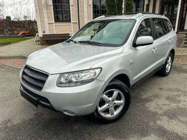 Geniş pencereli bir binanın yakınındaki düz kaldırımda gümüş renkli bir SUV park halinde. Sahne yeşil ağaçlar ve açık gökyüzü olan bir yerleşim alanı..