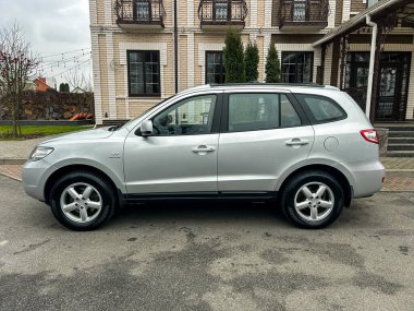 Balkonlu tuğla bir binanın yanına gümüş renkli bir SUV park edilmiş. Çevrede ağaçlar ve yapının net bir görüntüsü yer alıyor. Gökyüzü bulutlu ve bulutlu.