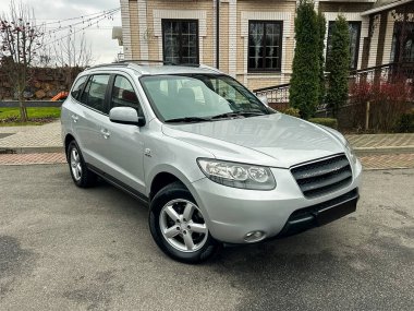 Bir binanın önünde park edilmiş gümüş renkli bir SUV görüldü. Bölgede bir taş patika ve çok sayıda yeşil ağaç yer alıyor. Gökyüzü bulutlu ve araç ileriye bakıyor..